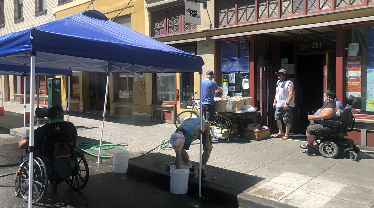 A canopy tent is on the sidewalk outside the Street Roots office. Someone sits under the shade of the tent and there are buckers of water and a hose running under the tent.There are three other people by the door of the office filling a table with supplie