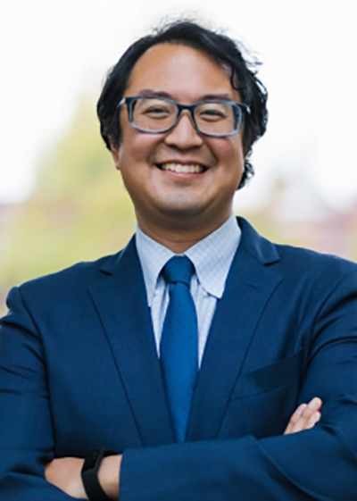Duncan Hwang smiles for a photo. He is wearing a suit and glasses and his arms crossed.
