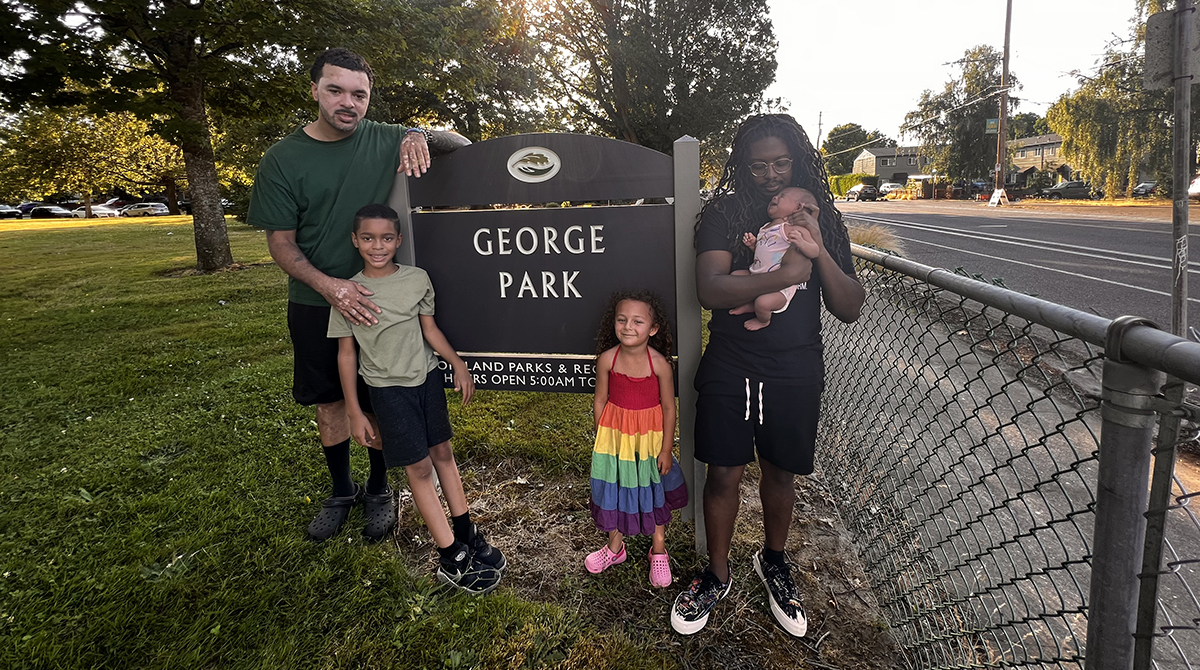 Mat Randol and Donovan Scribes stand on the side of a sign that says “George Park.” Donovan is holding his baby and Mat’s kids stand next to him in front of the sign.