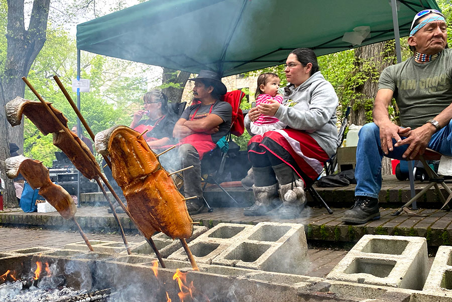 People sit around a small fire where salmon is being cooked on stakes.