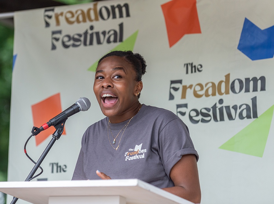 A woman speaks into a microphone at a podium. She is wearing a gray shirt that says "The Freadom Festival" and a backdrop behind her says the same.