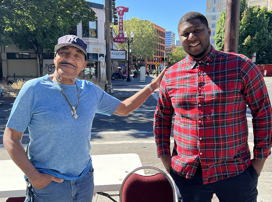 Desmond Hardigan smiles and has a hand on the shoulder of DeVon Pouncey. They are both smiling and standing on the sidewalk on a sunny day.