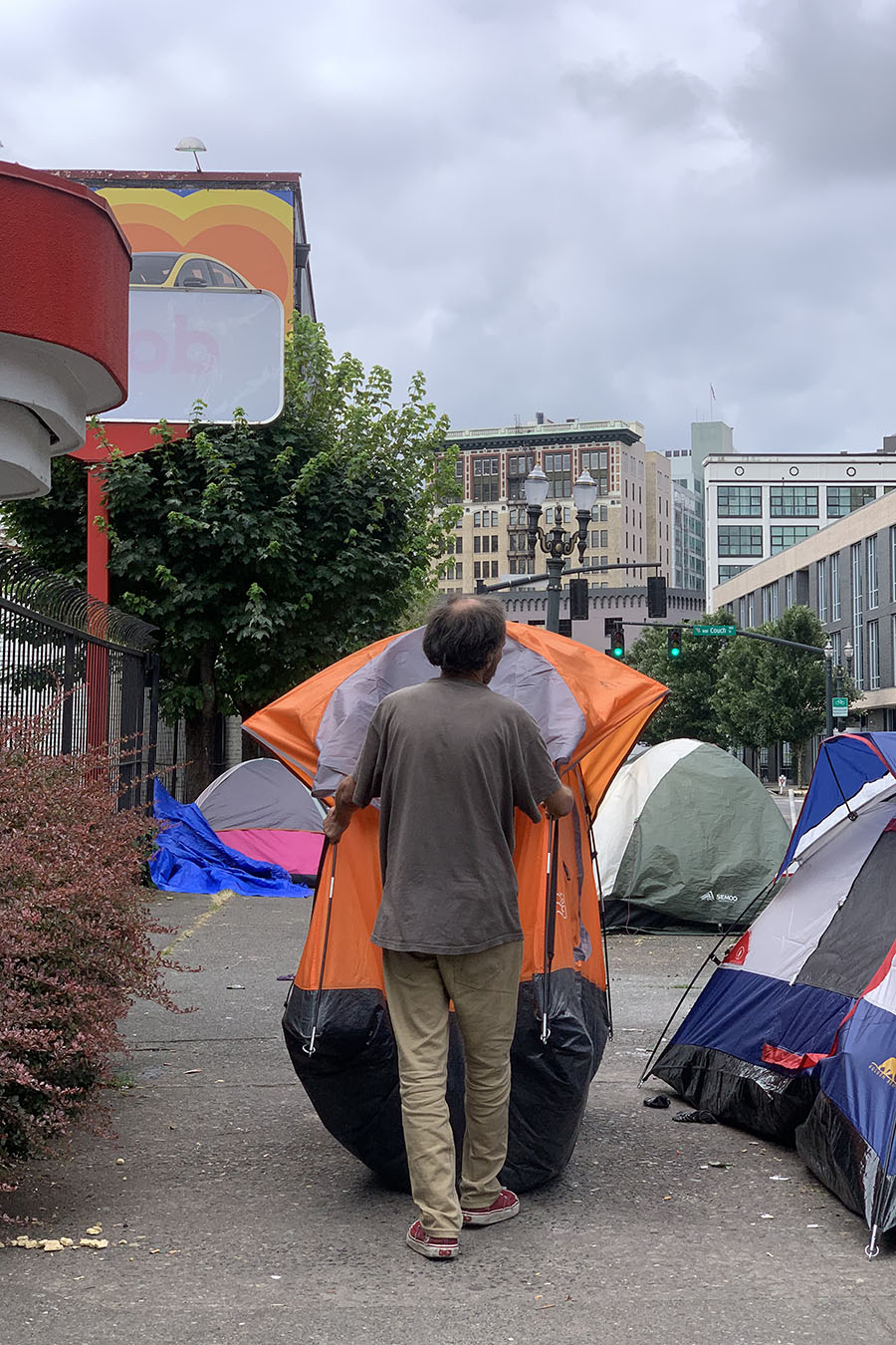 A view of a man from behind. He walks on the sidewalk holding a tent in his arms.