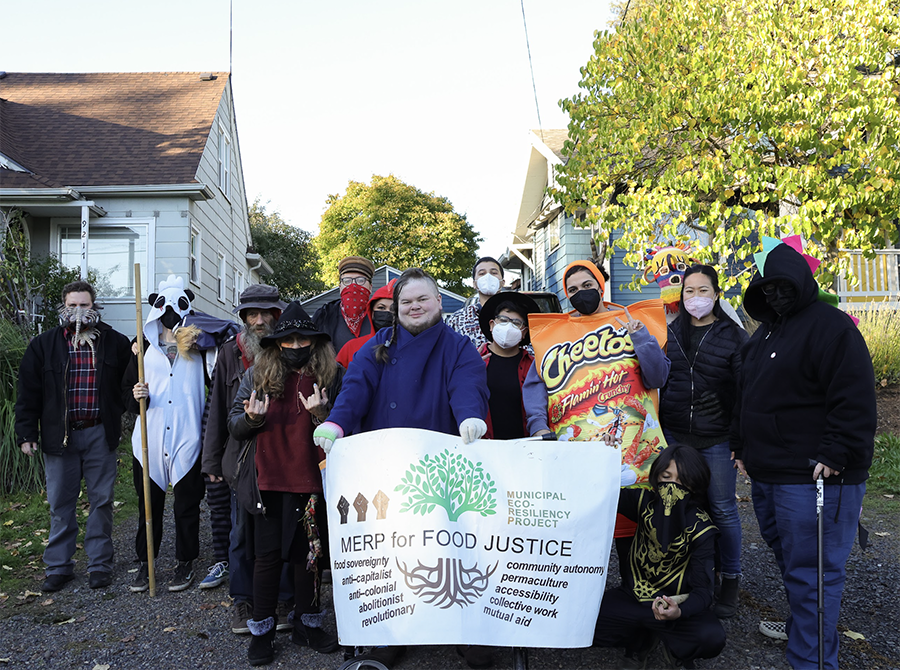 A group photo of people wearing Halloween costumes. The group stands between two houses and holds up a banner that says, "MERP FOR FOOD JUSTICE"