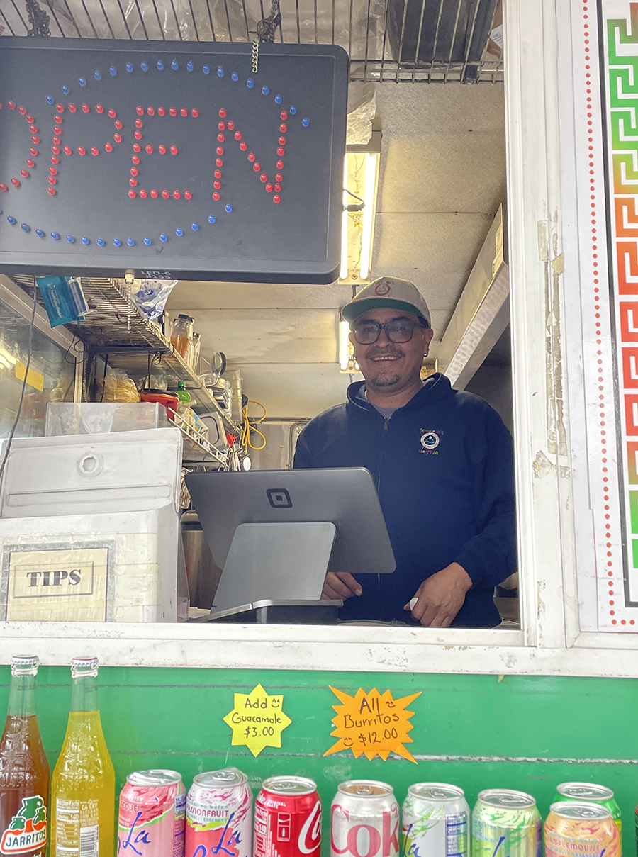 Fernando Rodriguez stands inside his food cart and smiles. He stands behind a register and below an "Open" sign.
