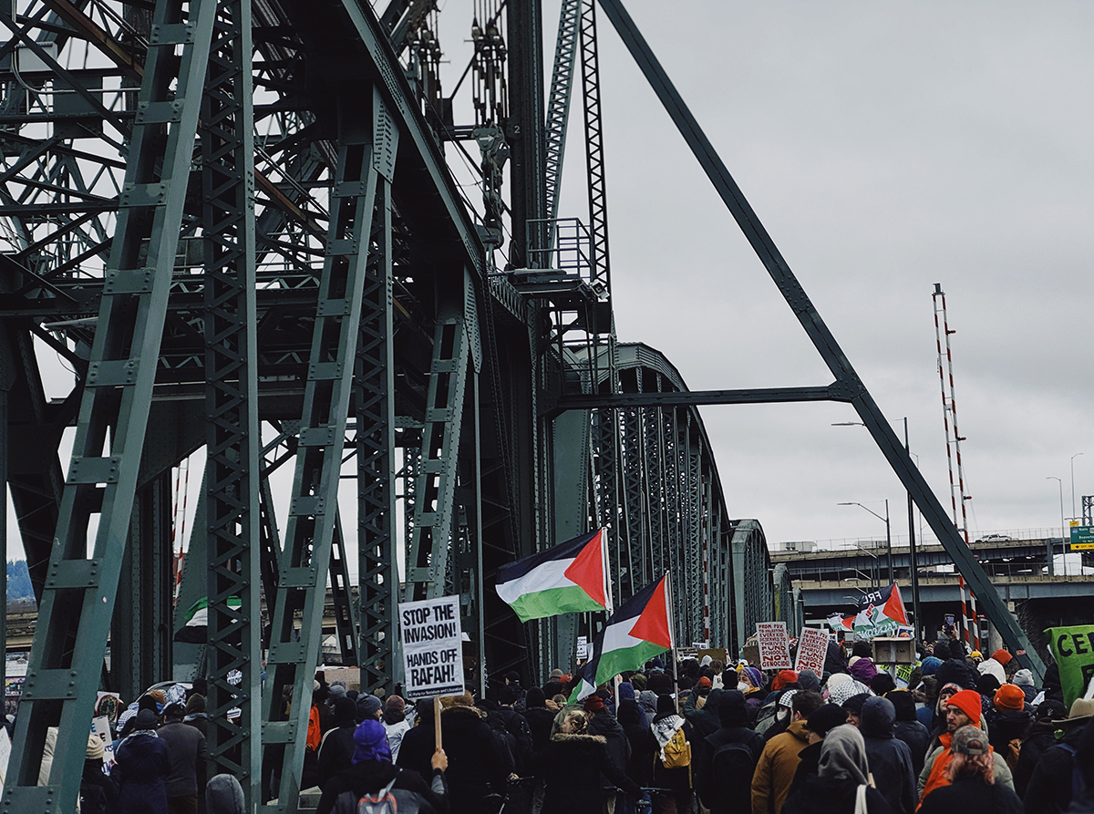 A rear view of a group of people marches across the Hawthorne Bridge. Many are holding up signs and Palestinian flags.