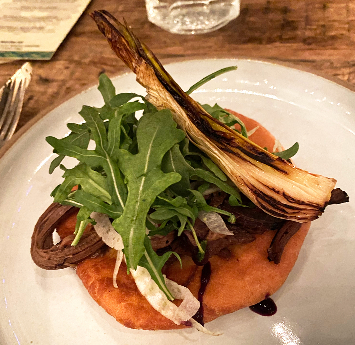 Frybread topped with wojapi berry sauce, braised elk, grilled wild onion and arugula is on a white ceramic plate