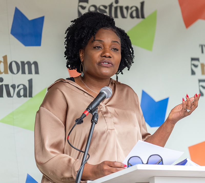 A woman is standing at a podium and speaking into a microphone with a backdrop behind her that says "The Freadom Festival."