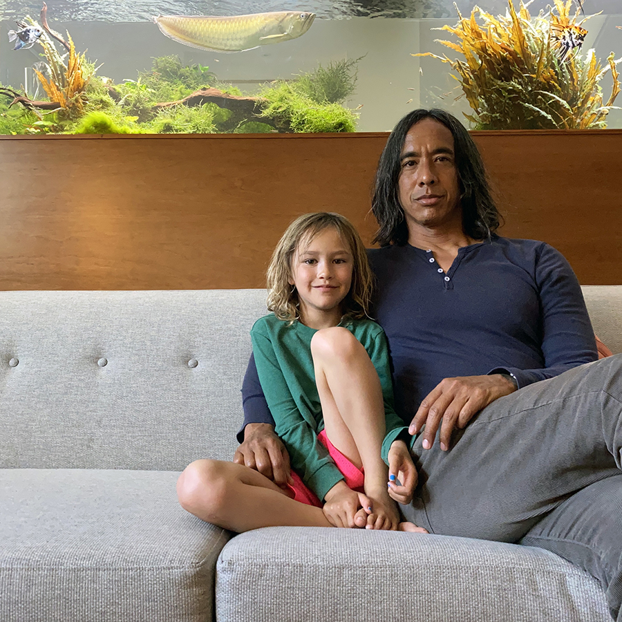 Artist Kip Fulbeck poses for a photo with his daughter sitting on a couch. Behind them is a tank with fish inside.