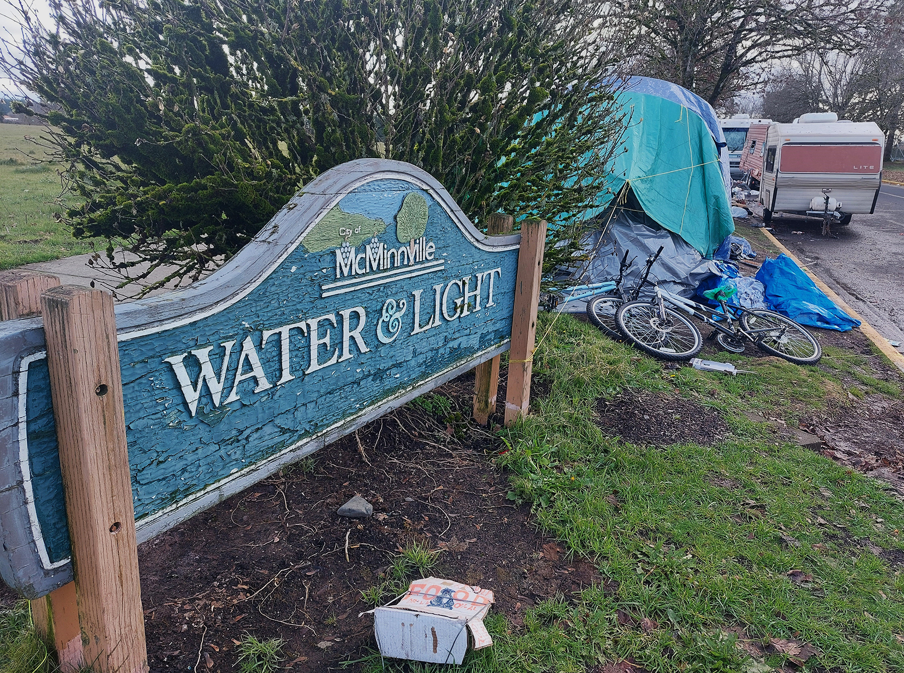 A wooden sign says "McMinnville water & light" is placed in grass next to some tents and a parked RV.
