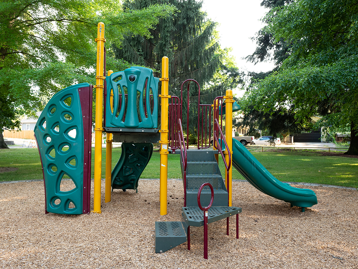 A play structure with two slides and a climbing wall sits in tan bark. The area is surrounded with trees.