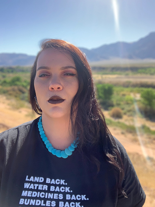Steph Littlebird poses for a photo with a black t-shirt with white text that says, "land back, water back, medicine back, bundles back."