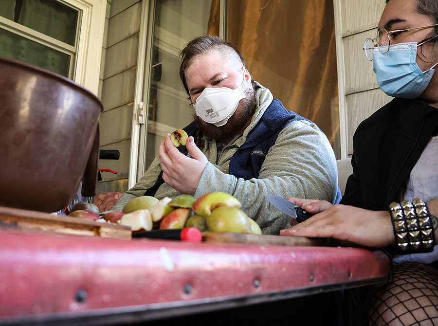 Two people sit at a table with a cutting board, knife and apples. one person is observing an apple slice in their hand.