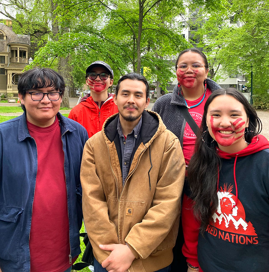 Five people stand together to pose for a group photo. One of them has their face decorated with a red handprint over their mouth.