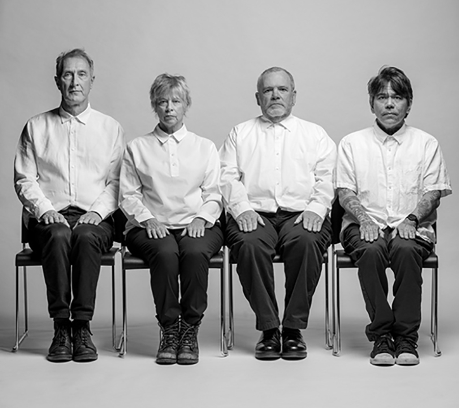 Jon King, Sara Lee, Hugo Burnham and David Pajo sit in a row in chairs wearing white button up shirts and black pants.