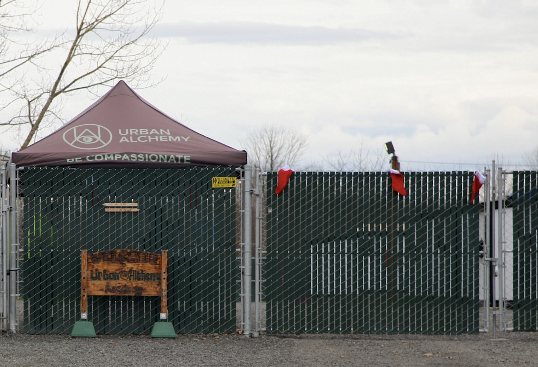 The exterior of the Reedway Safe Rest Village covered by a green fence. Behind the fence is a canopy tent that says "Urban Alchemy. Be compassionate."