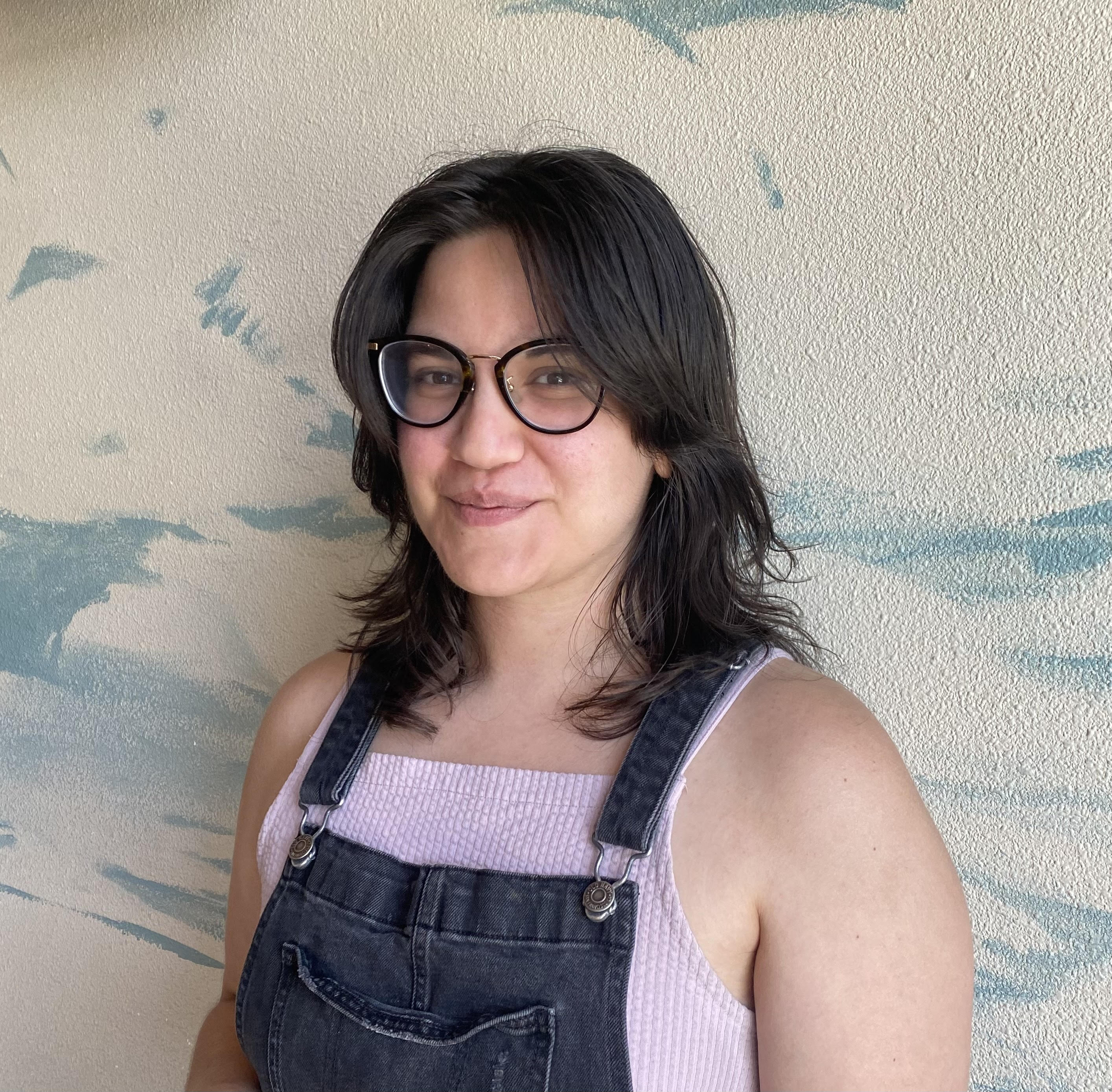 A portrait photo of Street Roots staff member Domenica Gavin. Domenica has half-long dark brown hair, loosely styled. She is wearing black-rimmed glasses and is looking into the camera at an angle. She is wearing a pink top with a pair of overall jeans 