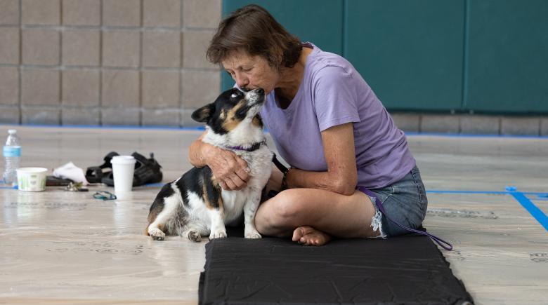 A person leans over and kisses her dog's head as the dog looks up at her. They are both sitting on a floor on a mat.