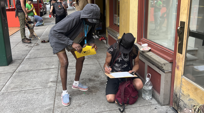 One man leans over as another crouches on the sidewalk, signing a petition on a clipboard