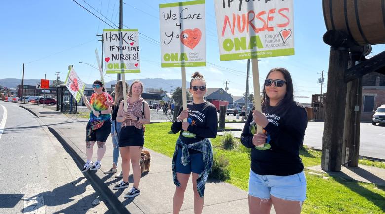 People stand in a line on a sidewalk on a sunny day. The people are holding up signs that say "honk if you love nurses."