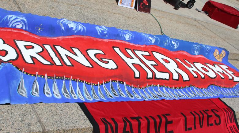 A photo of a banner that reads "bring her home" and another sign beneath that is cut off but reads, "Native Lives"