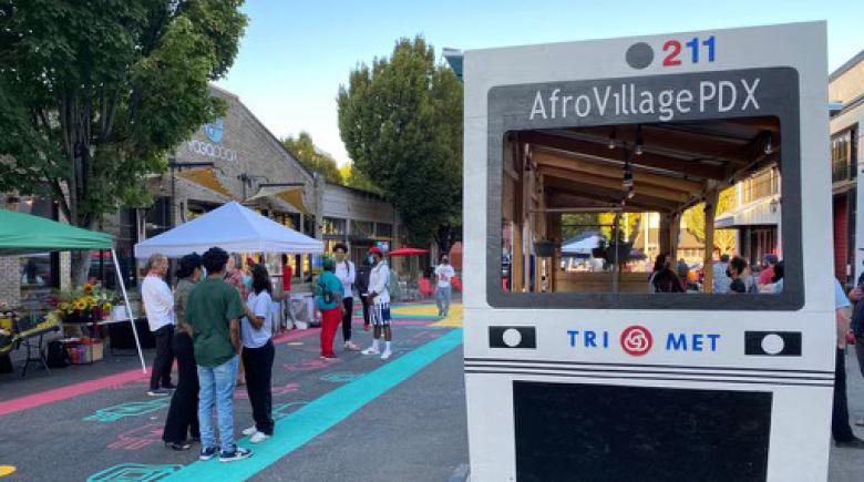 Photo of a street on the right is a construction of a MAX train that people can enter and it says "AfroVillage PDX." To the left are people talking in a group.