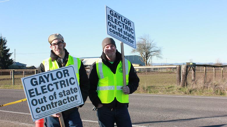 Two men hold signs that say "Gaylor electric out of state contractor" 