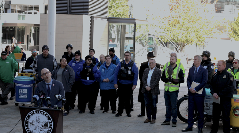 Andrew Hoan, Mayor Wheeler, Clean & Safe employees, and other elected officials stand in a crowd and listen as Hoan speaks at a podium.