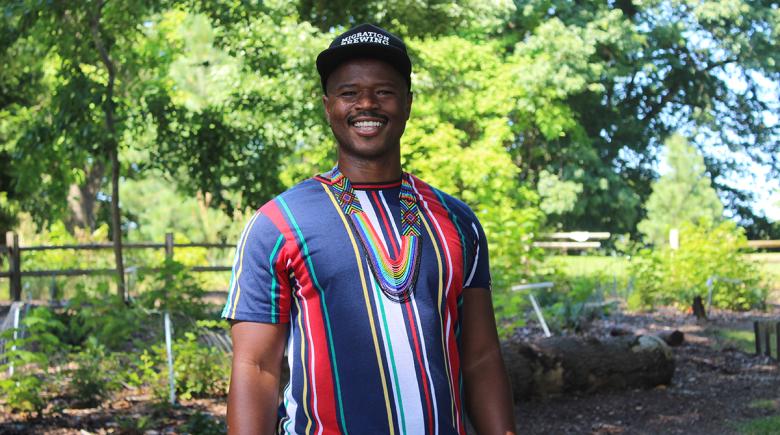Cameron Whitten poses and smiles for a photo. He is smiling and wearing a hat and a striped shirt. He is standing among greenery.