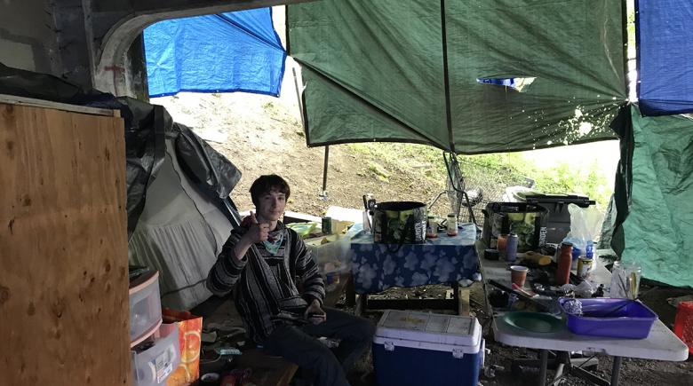 Austin sits on a bench, giving a thumbs-up signal, surrounded by folding tables, kitchen supplies and tarps.