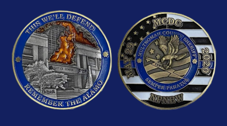 A photo of the front and back sides of the challenge coins.  The coin in question is emblazoned with an illustration of the Justice Center engulfed in flames on one side, encircled by the phrases “this we’ll defend,” and “remember the Alamo.” 