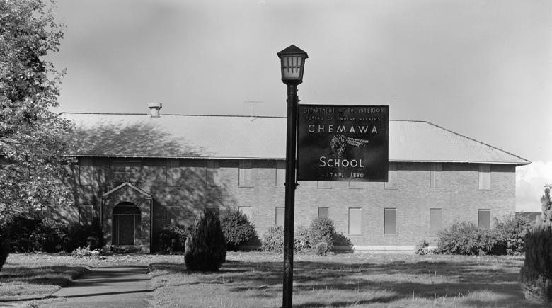 A sign says "Department of the Interior. Bureau of Indian Affairs. Chemawa Indian School Estab. 1880" in front of a building on the Chemawa Indian School campus.