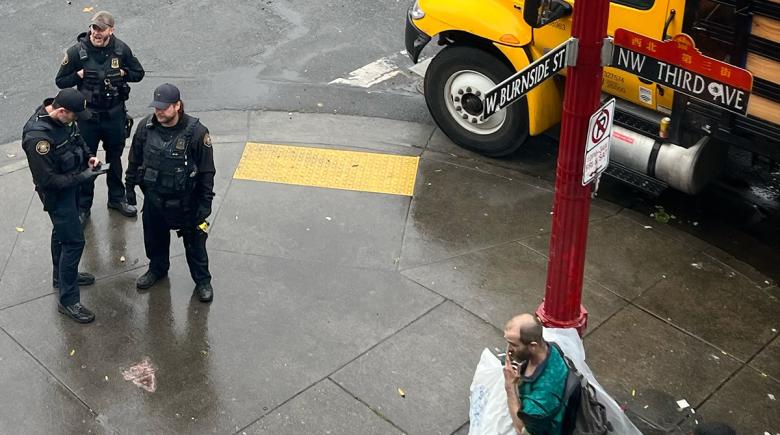 A street sign says "NW Third Ave" and the cross street sign says "W Burnside St." Below, on the sidewalk are three police officers and a man smoking is standing beneath the sign.