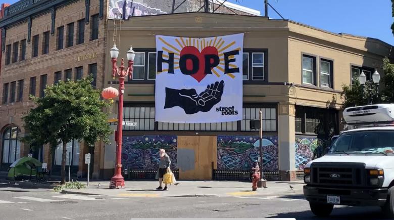 A photo of a building on West Burnside with a white hand-painted banner hanging over it. The banner says, "HOPE" and "Street Roots." The words sit above an open hand with yellow beams surrounding it.