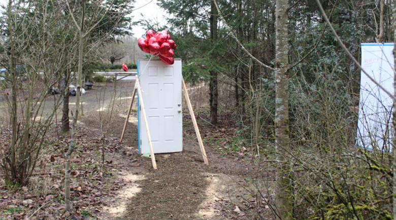A white door with balloons attached to it stands among trees on a dirt path.