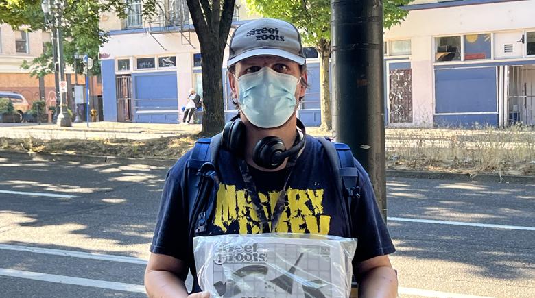 Jason stands for a photo outside on the sidewalk. He is wearing a Street Roots hat and holding a copy of the newspaper.
