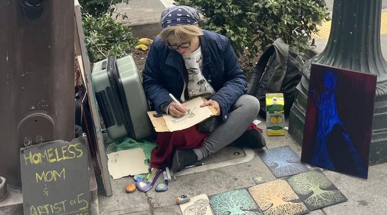 Karen Engelhardt sits on a sidewalk with a marker in hand, working on an art piece. To her left is a cardboard sign that says, "Homeless mom and artist of 5"