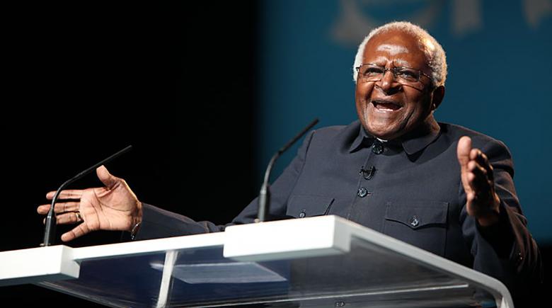 A photo of Archbishop Desmond Tutu in 2011 with his hands raised as he speaks standing over a podium. 
