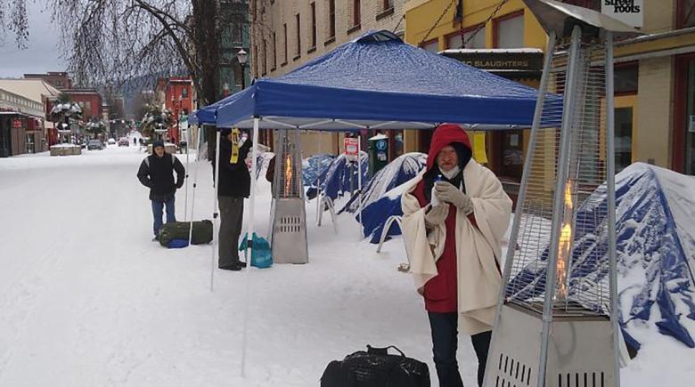 People outside street roots office with snow on the ground and space heaters. They are standing near the heaters and under the tents to keep warm during the snow storm. 