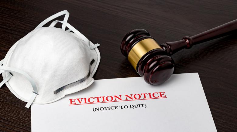A face covering/mask and a gavel sit on a wooden table with a piece of paper that says "Eviction Notice. Notice to quit" and a letter starts below that says, "Dear Tenant"