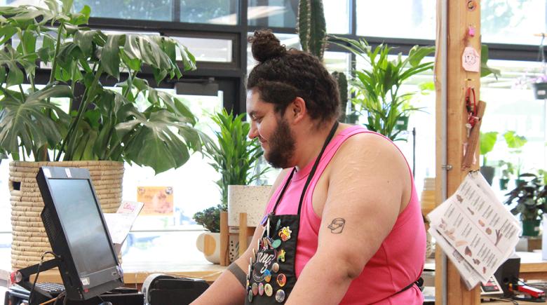 A person wearing a tank top and an apron covered in decorative pins works at the checkout line at Fang! and Salty's
