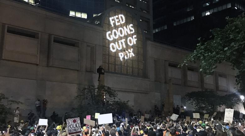 A message is projected onto a window, reading "Fed Goons Out of PDX"