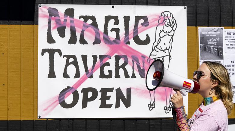 In the foreground a sign that says "Magic Tavern open" with an illustration of a woman has a pink "X" across the sign. To the right of the sign, a person holds a megaphone and is speaking into it.
