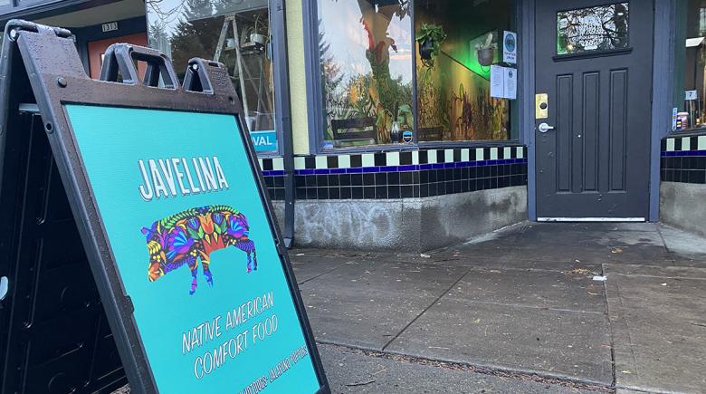 An A-frame sign sits on a sidewalk advertising Javelina. The sign says "Javelina. Native American Comfort food" and has a blue background and a rainbow colored Javelina.