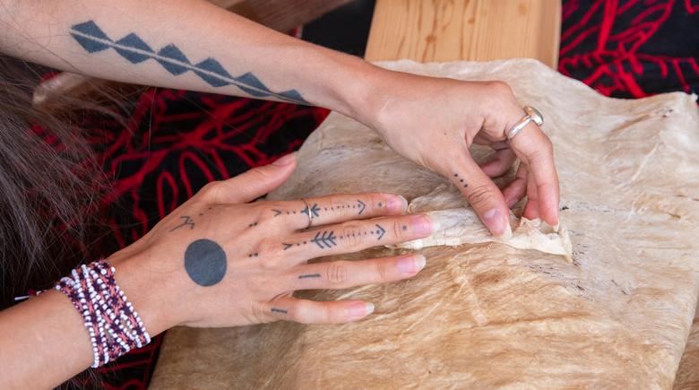 The photo is focused on Lehuauakea's hands. They are tattooed and wearing rings as they work with bark cloth.