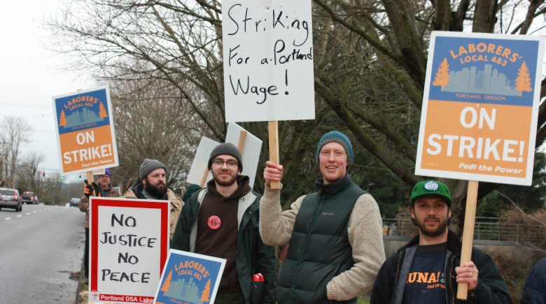 A group of people holding signs. One of the signs says, "striking for a Portland wage," "no justice no peace," and "on strike"