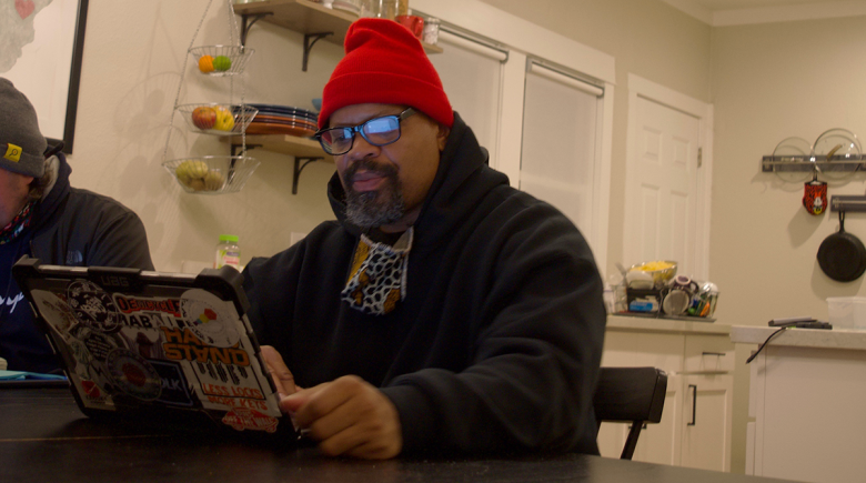 A photo of Mic Crenshaw with his laptop at a table in his kicthen. Mic wears a red beanie and black glasses. 