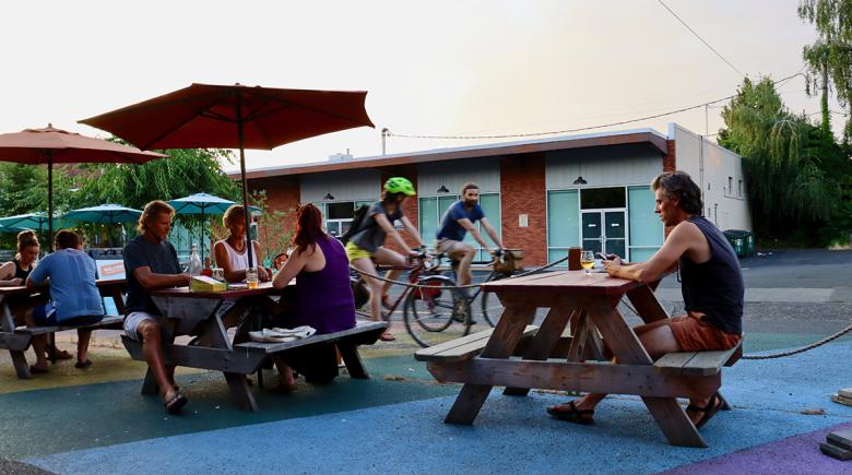 Cyclists bike down the street adjacent to an outdoor dining area while people are eating and drinking. The dining area has a rope separating it from the road and some of the tables have umbrellas.