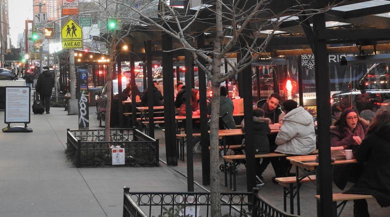 A photo of an outdoor dining structure adjacent to a city sidewalk. People are sitting and eating at the tables.