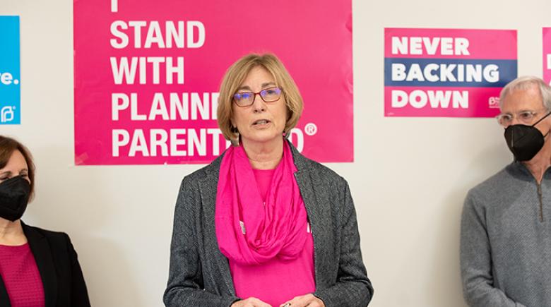 Anne Udall is speaking with two other people on the edges of the photo standing with face masks on. Anne wears glasses and stands in front of a poster that reads "Stand with planned parenthood"